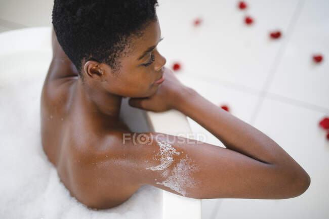 Sonriente mujer afroamericana en baño relajante en baño de espuma. estilo de vida doméstico, disfrutando del tiempo libre de autocuidado en casa. - foto de stock