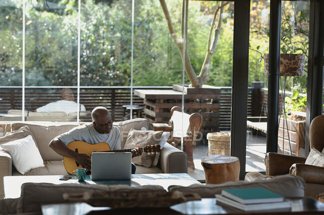 Feliz hombre afroamericano senior en la soleada sala de estar moderna tocando la guitarra. estilo de vida de jubilación, pasar tiempo solo en casa. - foto de stock