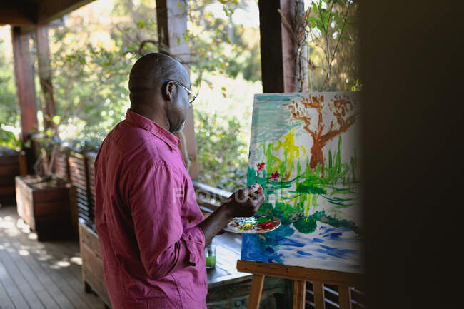Hombre afroamericano mayor en la pintura de balcón soleado. estilo de vida de jubilación, pasar tiempo solo en casa. - foto de stock