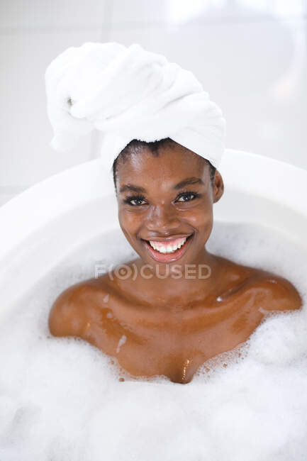 Портрет улыбающейся африканской американки в ванной комнате, расслабляющей в ванной, смотрящей в камеру. домашний образ жизни, наслаждаясь отдыхом на дому. — стоковое фото