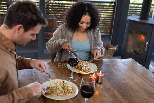 Happy diverse couple in living room sitting at table eating dinner together. spending time off at home in modern apartment. — Stock Photo