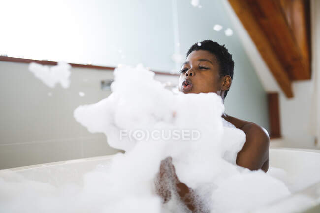 Щаслива афро-американська жінка відпочиває у ванній, розважаючись, пускаючи піну для ванн. Домашній спосіб життя, дозвілля для себе вдома. — стокове фото