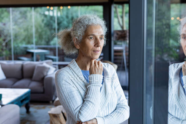 Nachdenkliche Seniorin im Wohnzimmer, die zum Fenster blickt. Lebensstil im Ruhestand, Zeit allein zu Hause verbringen. — Stockfoto