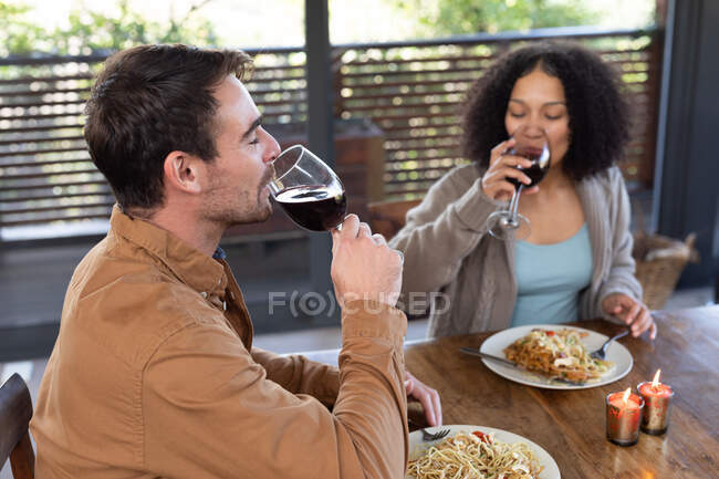 Feliz pareja diversa en la sala de estar sentados en la mesa cenando juntos y bebiendo vino. pasar tiempo libre en casa en apartamento moderno. - foto de stock