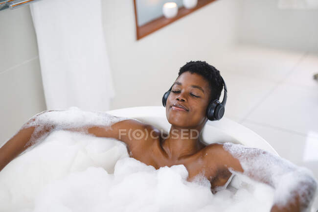 Femme afro-américaine souriante dans la salle de bain relaxante dans un bain en mousse portant un casque avec les yeux fermés. — Photo de stock