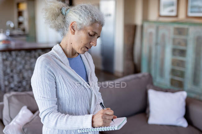 Eine ältere kaukasische Frau im Wohnzimmer steht und macht sich Notizen. Lebensstil im Ruhestand, Zeit allein zu Hause verbringen. — Stockfoto