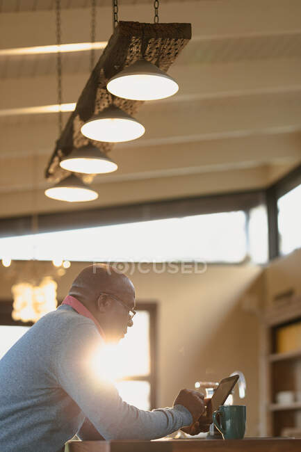 Senior afroamericano hombre situado en la moderna cocina soleada y el uso de la tableta. estilo de vida de jubilación, pasar tiempo solo en casa. - foto de stock