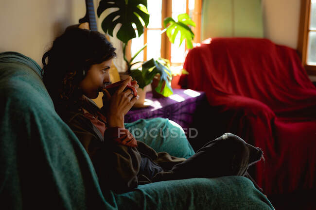 Mujer de raza mixta sentada y tomando café en la sala de estar soleada. estilo de vida saludable, disfrutando del tiempo libre en casa. - foto de stock