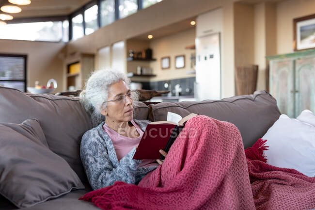 Mulher caucasiana sênior sentada no sofá e lendo livro na sala de estar moderna. estilo de vida aposentadoria, passar o tempo sozinho em casa. — Fotografia de Stock