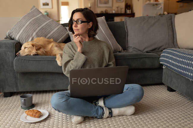 Femme caucasienne dans le salon avec son chien de compagnie, assise sur le sol, travaillant à l'aide d'un ordinateur portable. mode de vie domestique, profiter du temps libre à la maison. — Photo de stock