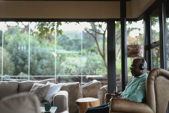 Détente homme afro-américain senior en utilisant des écouteurs dans le salon moderne. mode de vie à la retraite, passer du temps seul à la maison. — Photo de stock