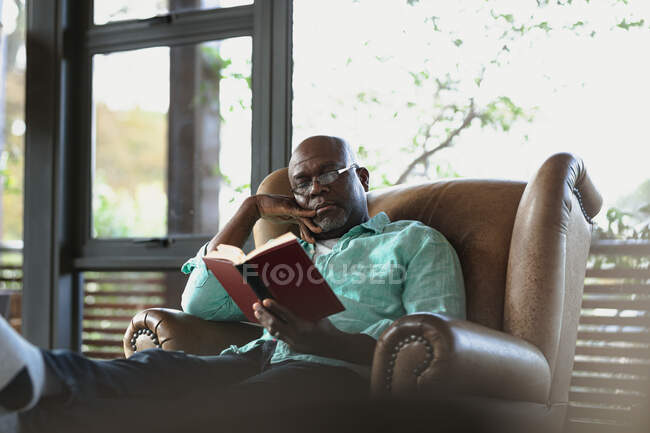 Homme afro-américain âgé assis sur le fauteuil et le livre de lecture dans le salon moderne. mode de vie à la retraite, passer du temps seul à la maison. — Photo de stock