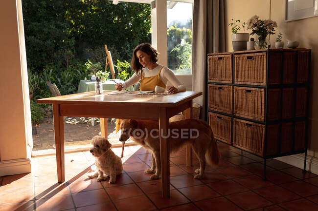 Mujer caucásica en la sala de estar con sus perros de compañía, sentada a la mesa de pintura. estilo de vida doméstico, disfrutando del tiempo libre en casa. - foto de stock