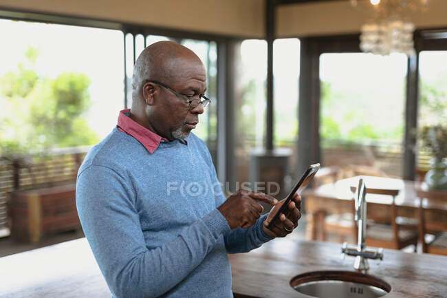 Hombre afroamericano mayor de pie y el uso de tableta en la cocina moderna. estilo de vida de jubilación, pasar tiempo solo en casa. - foto de stock