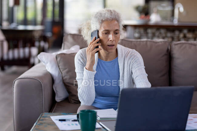 Mulher caucasiana sênior na sala de estar sentada no sofá, usando smartphone e laptop. estilo de vida aposentadoria, passar o tempo sozinho em casa. — Fotografia de Stock
