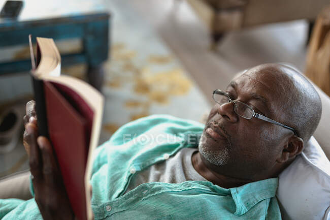 Relajante hombre afroamericano mayor acostado en el sofá y leyendo un libro. estilo de vida de jubilación, pasar tiempo solo en casa. - foto de stock