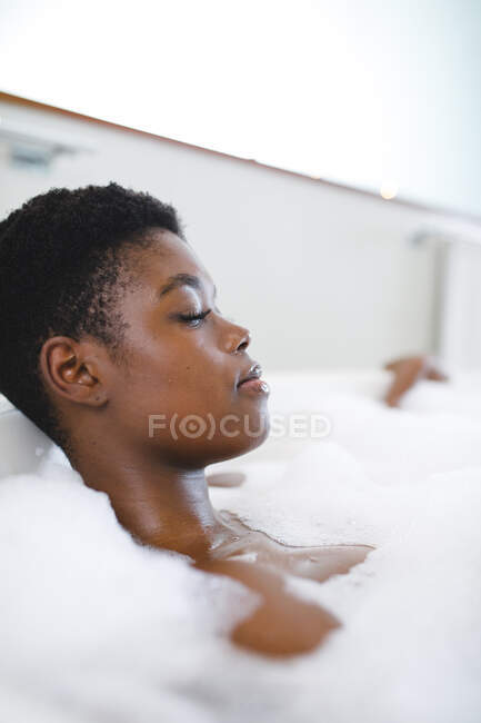 Mujer afroamericana sonriente en el baño, relajante en el baño con los ojos cerrados. estilo de vida doméstico, disfrutando del tiempo libre de autocuidado en casa. - foto de stock