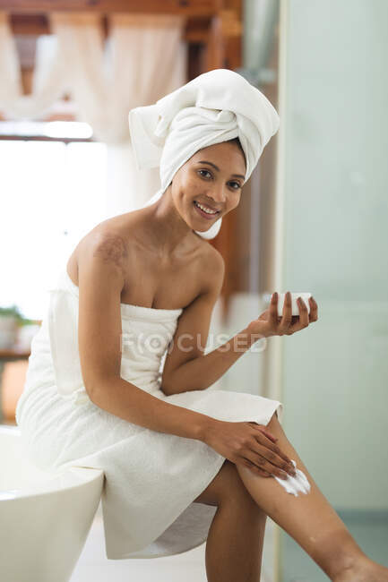Portrait of smiling mixed race woman in bathroom applying body cream to her legs. domestic lifestyle, enjoying self care leisure time at home. — Stock Photo