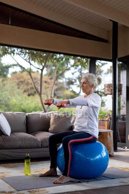 Femme caucasienne âgée dans le salon faisant de l'exercice, assise sur des haltères de levage de boules suisses. mode de vie à la retraite, passer du temps seul à la maison. — Photo de stock
