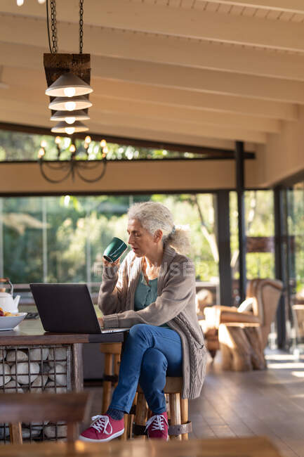 Расслабляет пожилую кавказку на кухне с помощью ноутбука и пьет кофе. пенсионного образа жизни, проводить время в одиночестве на дому. — стоковое фото