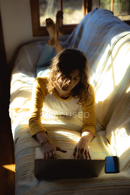 Mixed race woman lying and using laptop in sunny living room. healthy lifestyle, enjoying leisure time at home. — Stock Photo