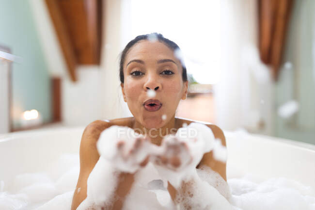 Porträt einer Frau mit gemischter Rasse in der Badewanne, die Spaß daran hat, Badeschaum zu pusten. häuslicher Lebensstil, selbstgepflegte Freizeit zu Hause genießen. — Stockfoto