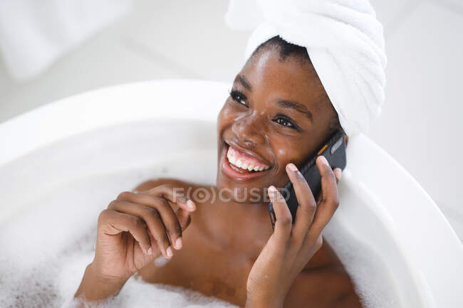 Lächelnde Afroamerikanerin im Badezimmer, entspannt in der Badewanne im Smartphone-Gespräch. häuslicher Lebensstil, selbstgepflegte Freizeit zu Hause genießen. — Stockfoto