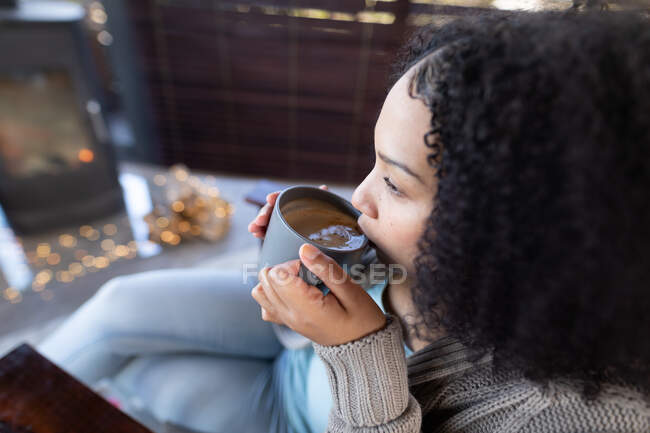 Feliz mujer de raza mixta en la sala de estar sentado junto a la chimenea sosteniendo taza y beber café. pasar tiempo libre en casa. . - foto de stock
