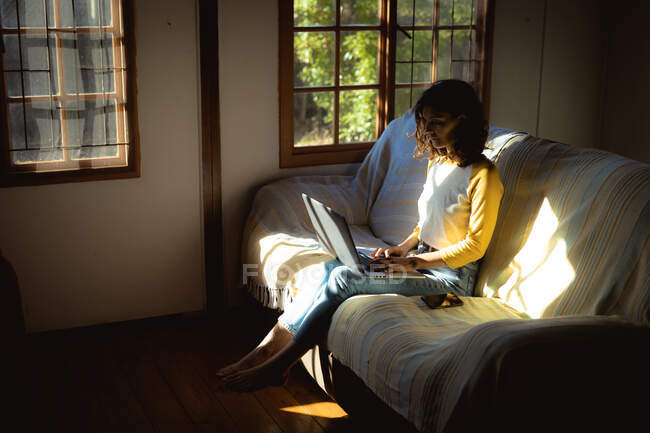 Змішана расова жінка сидить і використовує ноутбук у сонячній вітальні. здоровий спосіб життя, насолоджуючись дозвіллям вдома . — стокове фото
