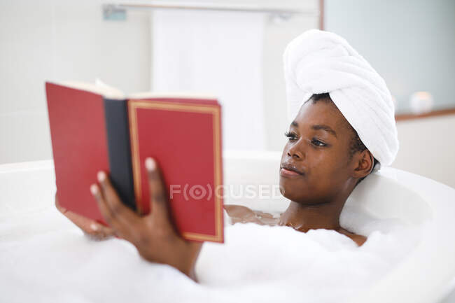 Mujer afroamericana feliz en baño relajante en libro de lectura de baño. estilo de vida doméstico, disfrutando del tiempo libre de autocuidado en casa. - foto de stock