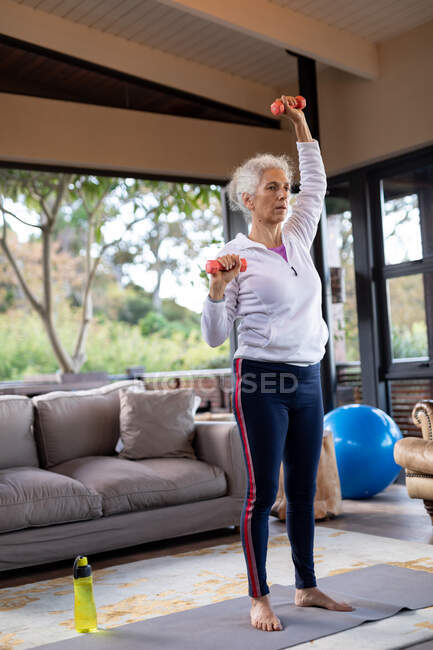 Femme caucasienne âgée dans le salon faisant de l'exercice, debout et soulevant des haltères. mode de vie à la retraite, passer du temps seul à la maison. — Photo de stock