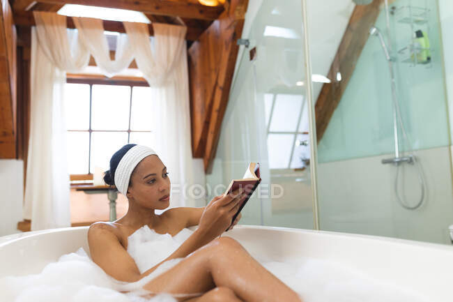 Mixed race woman in bathroom, relaxing in bath reading book. domestic lifestyle, enjoying self care leisure time at home. — Stock Photo