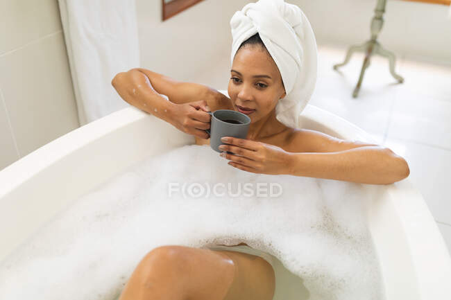 Glückliche Mischlingsfrau im Badezimmer, die eine Badewanne hat und Kaffee trinkt. häuslicher Lebensstil, selbstgepflegte Freizeit zu Hause genießen. — Stockfoto