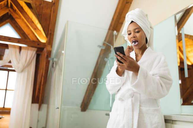 Mixed race woman in bathroom using smartphone and brushing her teeth. domestic lifestyle, enjoying self care leisure time at home. — Stock Photo
