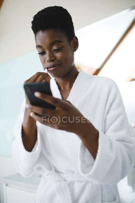 Africano mulher americana em casa de banho escovar os dentes e usando smartphone. estilo de vida doméstico, desfrutando de tempo de lazer auto-cuidado em casa. — Fotografia de Stock