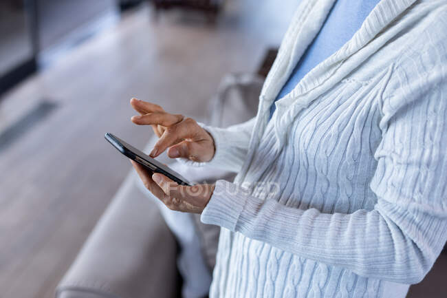 Seniorin im mittleren Lebensalter im Wohnzimmer stehend und mit Handy. Lebensstil im Ruhestand, Zeit allein zu Hause. — Stockfoto