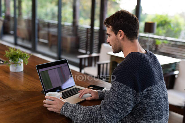 Hombre caucásico sentado en la mesa en la cocina trabajando remotamente con el ordenador portátil. en casa en apartamento moderno. - foto de stock