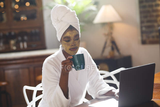 Sorrindo mulher americana africana com máscara facial de beleza na sala de estar usando laptop e beber café. — Fotografia de Stock