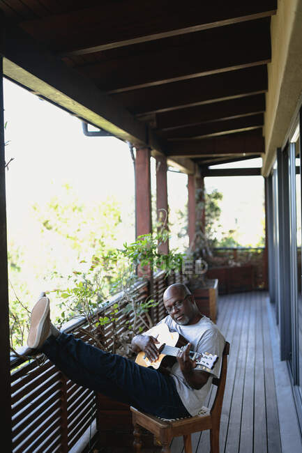 Feliz homem afro-americano sénior na varanda ensolarada a tocar guitarra. estilo de vida aposentadoria, passar o tempo sozinho em casa. — Fotografia de Stock