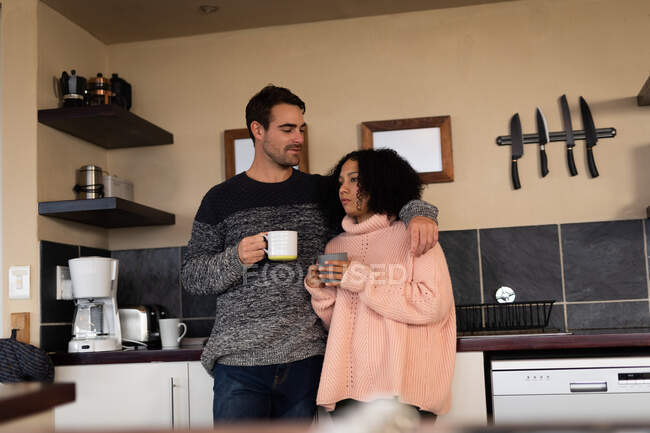 Feliz pareja diversa en la cocina abrazando beber café y sonreír. pasar tiempo libre en casa en apartamento moderno. - foto de stock