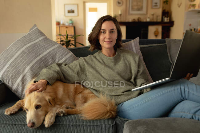 Портрет усміхненої білої жінки у вітальні, сидячи на дивані зі своїм домашнім собакою, використовуючи ноутбук. домашній спосіб життя, насолоджуючись дозвіллям вдома . — стокове фото