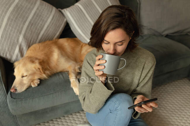 Caucasian woman in living room with her pet dog, using smartphone and drinking coffee. domestic lifestyle, enjoying leisure time at home. — Stock Photo