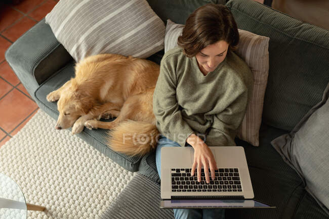 Mujer caucásica en la sala de estar, sentada en el sofá con su perro mascota, utilizando el ordenador portátil. estilo de vida doméstico, disfrutando del tiempo libre en casa. - foto de stock