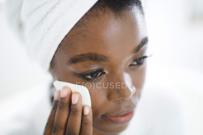 Улыбающаяся африканская американка в ванной очищает лицо хлопчатобумажной салфеткой для ухода за кожей. домашний образ жизни, наслаждаясь отдыхом на дому. — стоковое фото