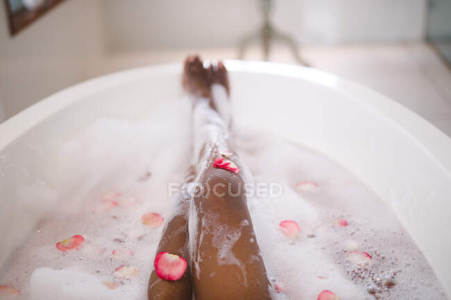 Sección baja de mujer afroamericana relajándose en un baño de espuma mimada con pétalos de rosa. estilo de vida doméstico, disfrutando del tiempo libre de autocuidado en casa. - foto de stock