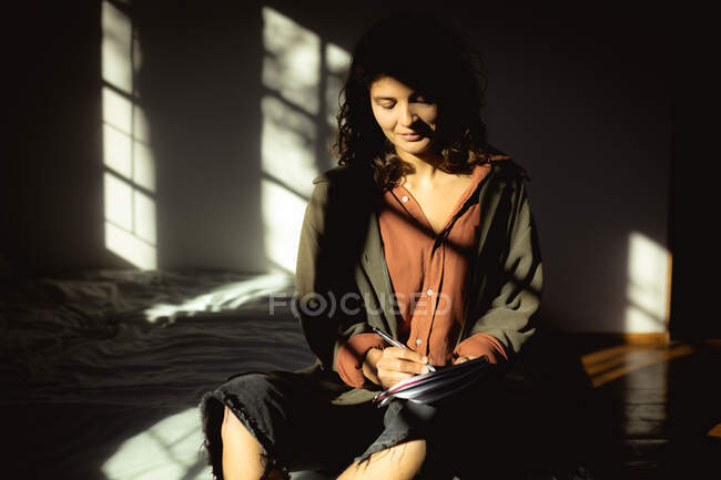 Mujer de raza mixta sentada y tomando notas en el dormitorio soleado. estilo de vida saludable, disfrutando del tiempo libre en casa. - foto de stock