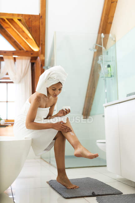 Mulher de raça mista no banheiro aplicando creme corporal em suas pernas. estilo de vida doméstico, desfrutando de tempo de lazer auto-cuidado em casa. — Fotografia de Stock