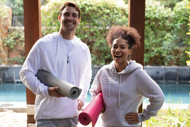 Retrato de feliz pareja diversa sosteniendo colchonetas de yoga antes de practicar yoga. pasar tiempo libre en casa. - foto de stock