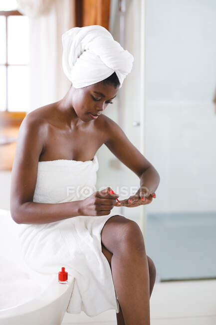 Sorrindo afro-americana na casa de banho pintando as unhas com esmalte vermelho. estilo de vida doméstico, desfrutando de tempo de lazer auto-cuidado em casa. — Fotografia de Stock
