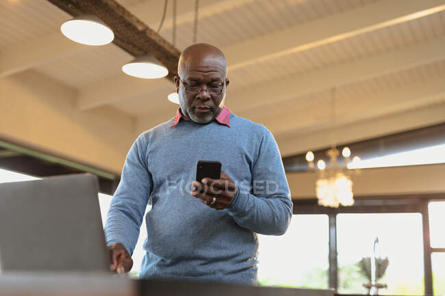 Hombre afroamericano mayor de pie y el uso de teléfonos inteligentes en la sala de estar moderna. estilo de vida de jubilación, pasar tiempo solo en casa. - foto de stock